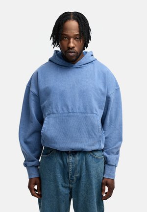 Blue hoodie made of soft fabric, featuring a front pouch pocket and ribbed cuffs. Paired with loose-fitting blue jeans.