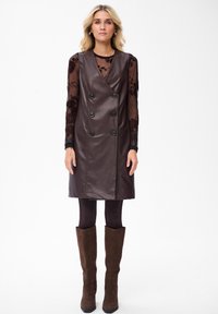 Kobieta ubrana w bezrękawnikową brązową skórzaną sukienkę na czarnym, kwiecistym, przezroczystym topie z długim rękawem, ciemne legginsy i brązowe zamszowe kozaki za kolano.