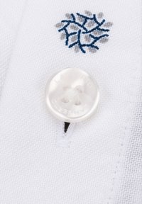 Biały materiał z teksturowanym splotem, z przezroczystym guzikiem i niebieskim haftowanym wzorem śnieżynki nad dziurką na guzik.