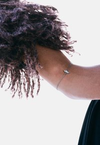 Braccialetto a catena d'argento con un piccolo ciondolo triangolare, indossato su un braccio nudo parzialmente coperto da capelli ricci. Lo sfondo è bianco uniforme.