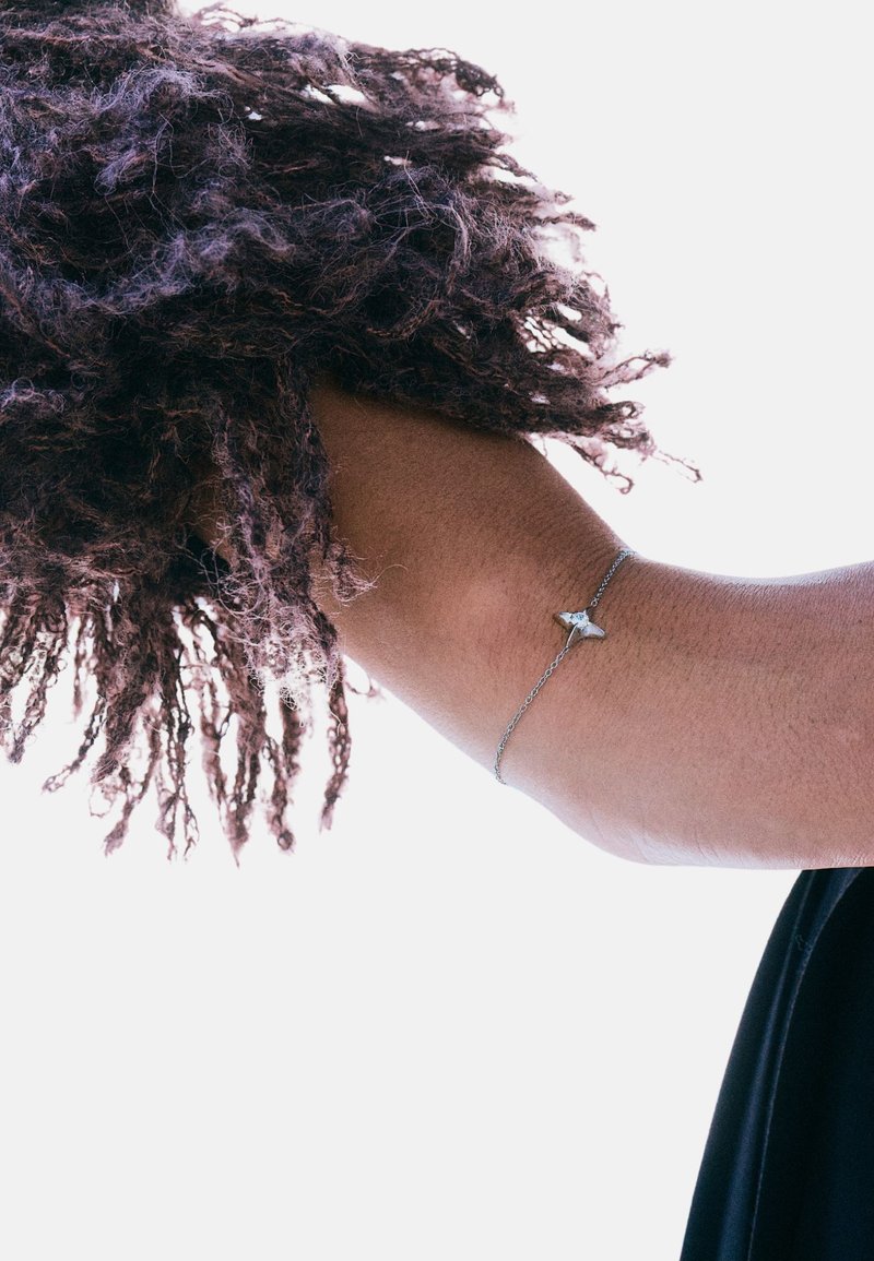 Braccialetto a catena d'argento con un piccolo ciondolo triangolare, indossato su un braccio nudo parzialmente coperto da capelli ricci. Lo sfondo è bianco uniforme.