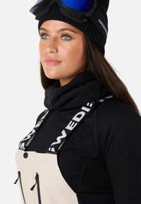 Sorte skitøj med ribbet tekstur, der har en cremet bib, sorte seler mærket "SVERIGE", og en sort hue med skibriller.