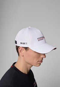Biała czapka baseballowa z czerwonymi akcentami, z logo "Porsche Motorsport" i "Mobil 1". Strukturalny design z płaskim daszkiem i oddychającym materiałem.