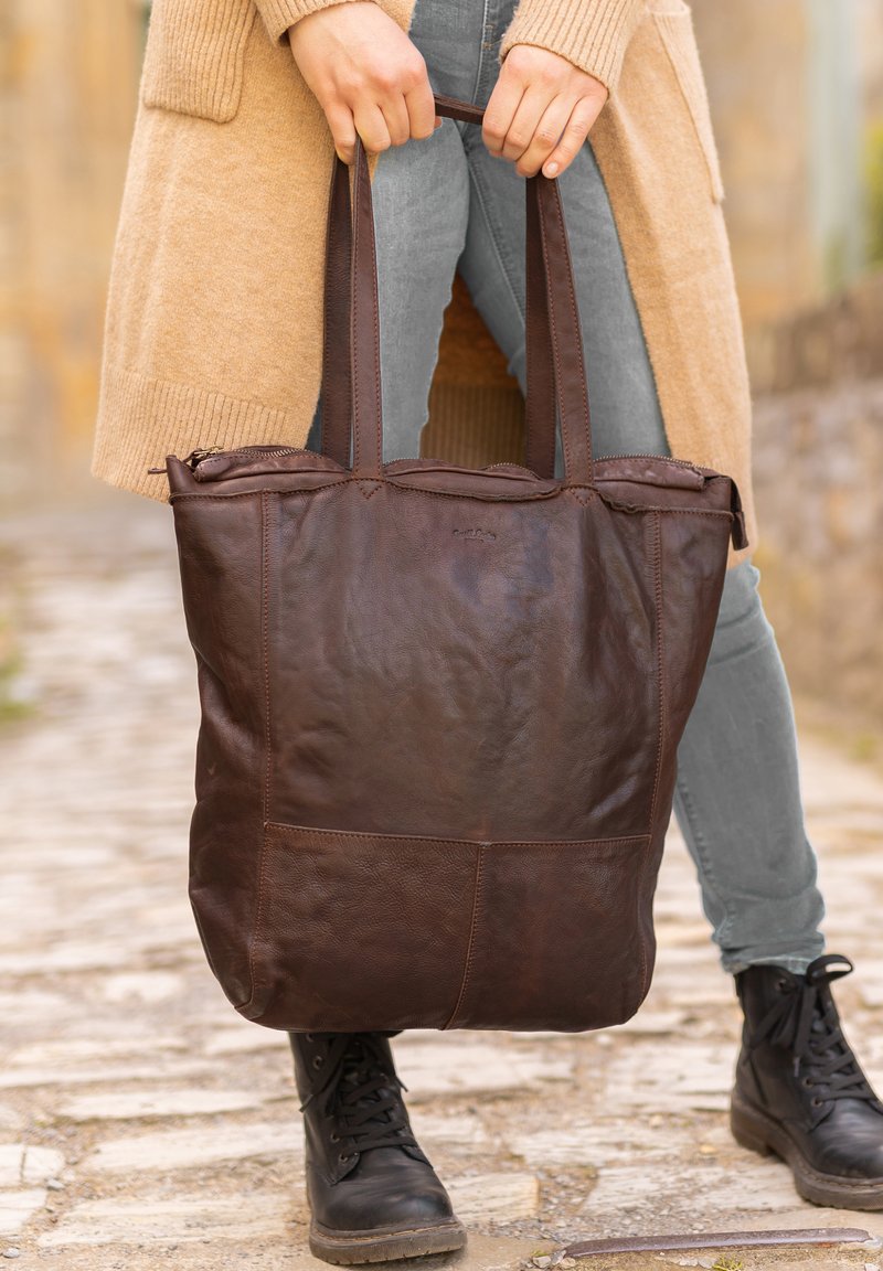 Brown leather tote bag with two long handles, featuring a zip top and visible stitching, held by a person in gray jeans and black boots.