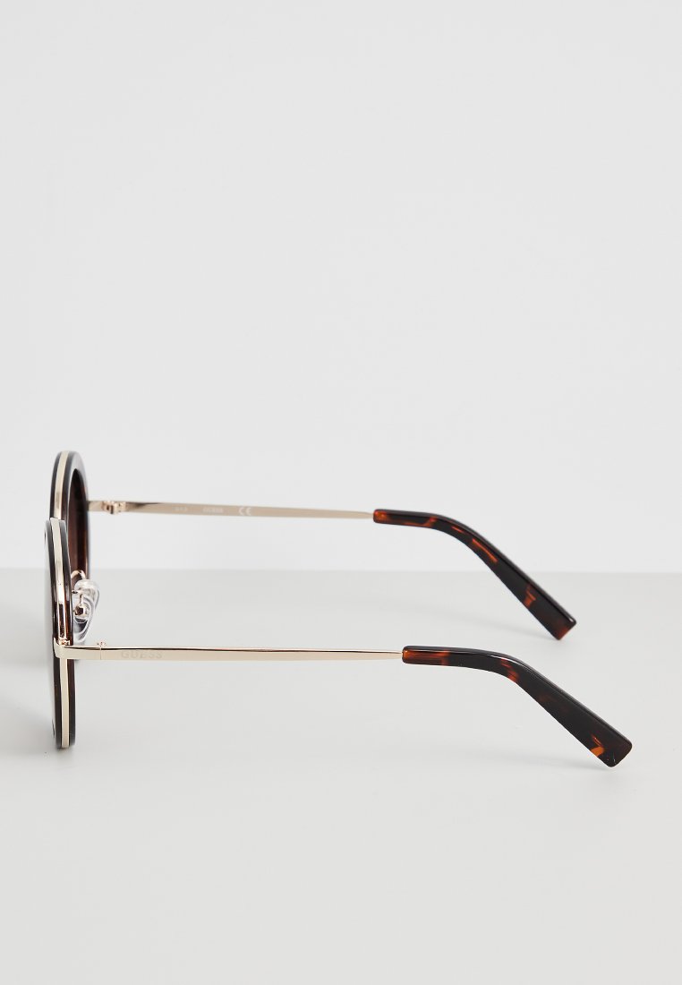 Occhiali da sole rotondi con sottili montature in metallo dorato e punte delle aste in plastica a motivo tartaruga su una superficie bianca.