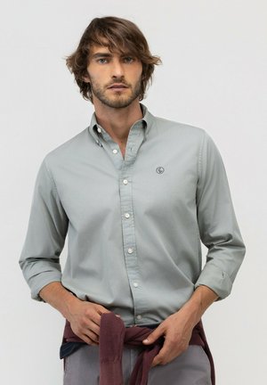 Hombre con cabello ondulado que lleva una camisa gris claro abotonada y sostiene un suéter morado oscuro atado alrededor de su cintura.