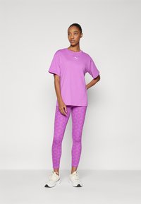 Lilla bomulds T-shirt med rund hals og hvidt logo, kombineret med lilla mønstrede leggings og hvide sportssko.