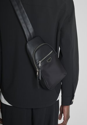 Bolso cruzado negro con dos cremalleras plateadas, llevado sobre la espalda de una persona con chaqueta y pantalones negros.