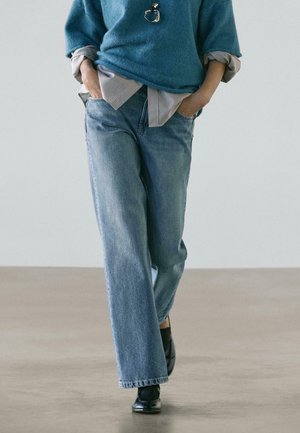 Persona che indossa jeans blu, scarpe nere, maglione blu sopra una camicia a righe, cammina con le mani in tasca su un pavimento e uno sfondo neutri.