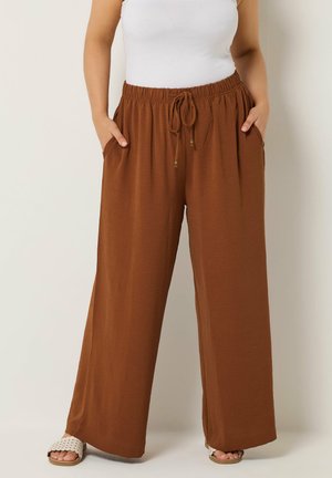 Kvinde iført brune bukser med vide ben, høj talje, snøre i taljen og sidelommer, kombineret med hvid top og hvide sandaler.