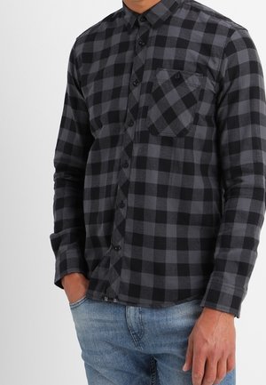 Mann trägt ein schwarz-grau kariertes Hemd mit Brusttasche, kombiniert mit hellblauen Jeans, steht mit einer Hand in der Hosentasche.