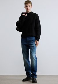 Mustast ribikootud kampsunist, sinistest sirge säärega teksadest, millel on heledam plekid, mustad jalanõud, õlal kandekott, tavalise tausta ees.