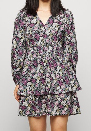 Frau trägt ein langärmliges, V-Ausschnitt, blumiges Kleid mit geraffter Taille und gestuftem Rock in lila, weißen und grünen Farbtönen.