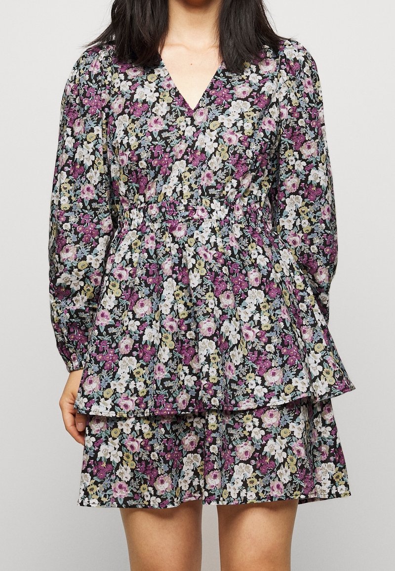 Kvinna klädd i en långärmad, V-ringad blommig klänning med samlad midja och en flikig kjol i lila, vitt och grönt.