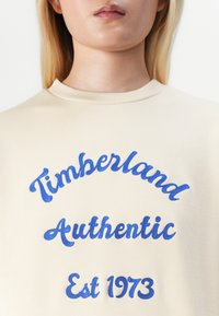 Hanorac bej cu text brodat albastru: "Timberland Authentic" și "Est 1973," având guler rotund și textură moale.