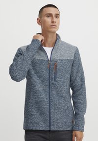 Mėlyna ir pilka zip-up striukė su tekstūruotu audiniu, kontrastingo panelių dizainu ir aukšta apykakle. Dėka rudo odinio užtrauktuko traukelio.