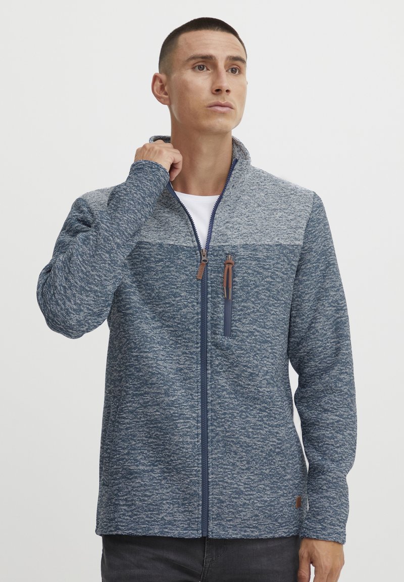 Mėlyna ir pilka zip-up striukė su tekstūruotu audiniu, kontrastingo panelių dizainu ir aukšta apykakle. Dėka rudo odinio užtrauktuko traukelio.