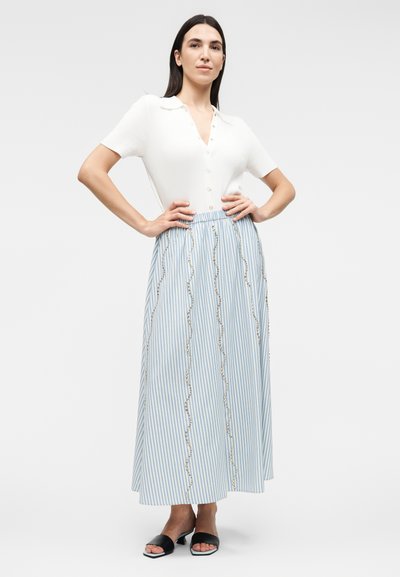 Γυναίκα όρθια με τα χέρια στη μέση, φοράει λευκό κοντομάνικο πουκάμισο με κουμπιά και μακριά μπλε και λευκή ριγέ φούστα με κυματιστά μοτίβα.
