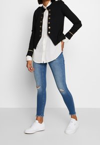 Chaqueta negra corta con botones dorados, llevada sobre una camisa blanca. Combinada con jeans ajustados rotos azules y zapatillas blancas.