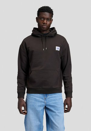 Homme debout devant un fond uni, portant un sweat à capuche Lee noir avec une poche avant et un jean bleu clair, face à l'objectif avec une expression neutre.