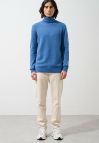 Světle modrý rolák vyrobený z měkké látky, spárovaný s béžovými kalhotami a bílými sportovními botami. Střih je volný s žebrovanými manžetami.