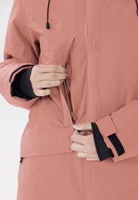 Veste imperméable rose avec une fermeture éclair à l'avant et des poignets à velcro. Caractérisée par un tissu doux et texturé, avec une tirette de fermeture éclair visible.