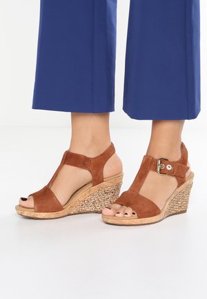 Des sandales à talons compensés en daim marron présentent un bout ouvert, un design en T et une texture tressée en synthétique sur le talon compensé, avec un accent à boucle.