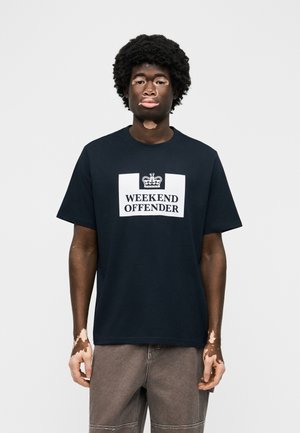 Νέος άνδρας με λεύκη, που φοράει μαύρο μπλουζάκι "Weekend Offender" και γκρι παντελόνι, στέκεται μπροστά σε απλό φωτεινό φόντο.