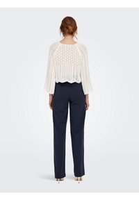 Haut blanc en tricot ajouré avec manches larges ; ourlet festonné. Porté avec un pantalon bleu marine et des talons. Fond en dégradé gris doux.