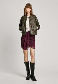 Olivově zelená kožená bomber bunda, bílý texturovaný svetr, burgundská květinová sukně, černé vysoké kozačky. Jednoduchý, moderní design.