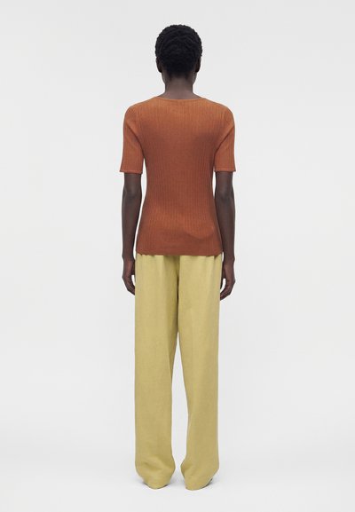 Άτομο με κοντά σγουρά μαλλιά στέκεται με την πλάτη προς τα εμπρός, φορώντας ριμπ πλεκτό τοπ με κοντά μανίκια σε σκουριά απόχρωση και φαρδιά μπεζ παντελόνια σε απλό φόντο.