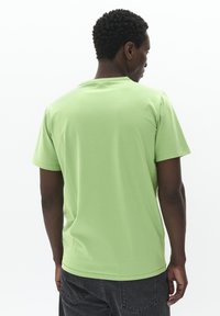 Camiseta de algodón verde con mangas cortas, cuello redondo y un diseño liso, vista desde la parte trasera. Llevada con pantalones gris oscuro.