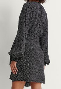 Robe noire à pois blancs, manches longues évasées, corsage ajusté, taille froncée, longueur au-dessus du genou, texture de tissu lisse.