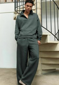 Conjunto de sudadera y pantalones a juego en gris carbón, con cuello con cremallera, corte holgado y textura de tela suave. Se presenta frente a un fondo de escalera.