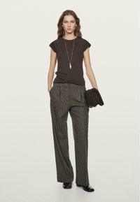 T-shirt aderente a maniche corte marrone, pantaloni a gamba larga grigi con vita alta, scarpe nere slim, tiene una pochette di tessuto scuro, collana con pendente d'oro.