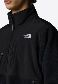 Black fleece jacket with a high collar, zip closure, and The North Face logo. Features textured material and front pockets with closures.