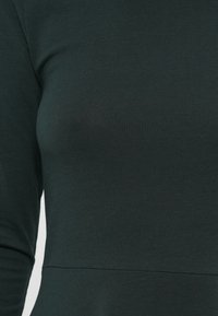 Haut à manches longues de couleur vert foncé, fabriqué en tissu doux et côtelé. Présente une coupe ajustée avec un détail de couture autour de la taille. Texture lisse.