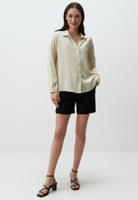 Beige langetermijn shirt met een subtiele bloemenstructuur, knoopsluiting aan de voorkant, gecombineerd met zwarte shorts en zwarte sandalen met bandjes.