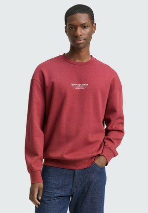 Bordeaux sweatshirt met geribbelde boorden en zoom, met een witte logo-print op de voorkant. Gekleed met blauwe spijkerbroeken.