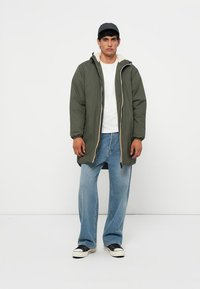 Veste matelassée vert olive avec une doublure de capuche beige, fermetures éclair, portée sur une chemise blanche et un jean bleu clair. Des baskets noires complètent le look.