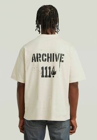 Crème T-shirt met grote zwarte tekst op de achterkant met de tekst "ARCHIVE 1114," korte mouwen en een ontspannen pasvorm.
