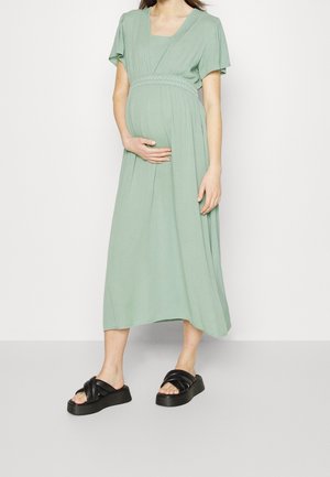 Kobieta w ciąży w jasnozielonej sukience midi z falbanami na rękawach i czarnych sandałach na platformie, stojąca na jednolitym jasnym tle.