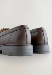 Brązowe skórzane mokasyny o teksturowanej powierzchni, kontrastowym przeszyciu i grubym czarnym gumowym podłożu. Okrągły design obcasa, bez widocznych ozdobników.
