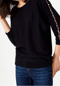 Svart tröja med trekvartslånga ärmar, featuring genomskinliga detaljer och vita knappar på sidan. Mjuk textur, avslappnad passform och ribbad fåll.