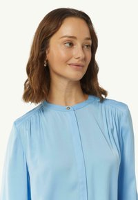 Jasnoniebieska bluzka z gładkiego materiału z okrągłym dekoltem, subtelnymi fałdami na ramionach i ozdobnym guzikiem przy kołnierzyku.