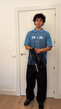 Mladý muž s kudrnatými vlasy, oblečený v modro pruhované tričko s nápisem "AIR" a černými kalhotami, drží složený tmavý oděv, stojí uvnitř vedle bílé dveře.