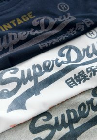 Naskládaná trička v námořnické modři, bílé a šedé. Každé má velké logo "SuperDry" v různých odstínech a jemnou bavlněnou texturu.