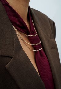 Brauner Blazer aus strukturiertem Stoff, mit einer tiefburgunderfarbenen Krawatte und drei goldenen Kettenakzenten, die über die Krawatte fallen.