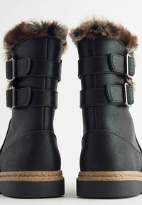 Schwarze Stiefel aus Kunstleder mit einem Kunstfellbesatz, zwei verstellbaren silbernen Schnallen und einer strukturierten Korksohle.