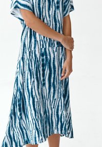 Vestido de patrón azul y blanco con mangas cortas y un diseño fluido. Presenta líneas verticales y tonos variados de azul. Textura de tela suave.